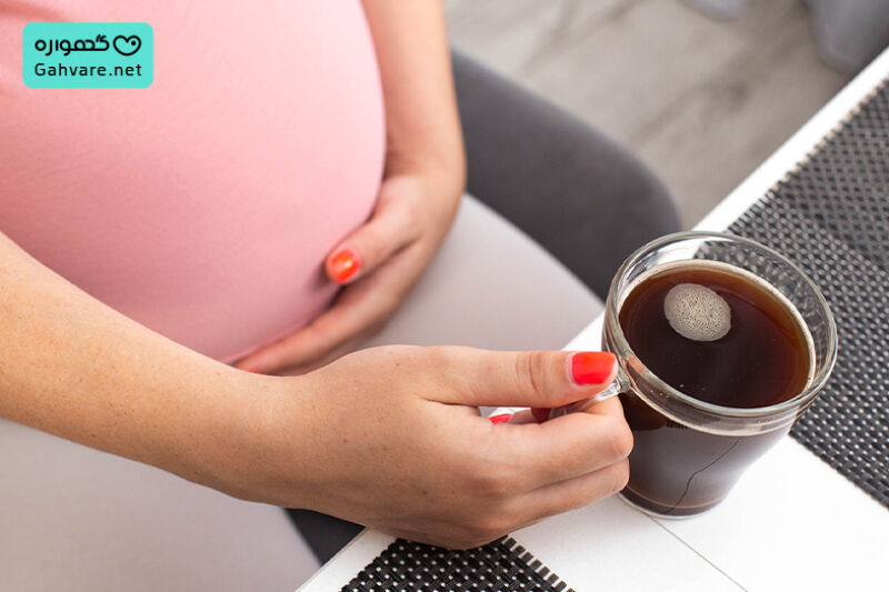 یک لیوان قهوه روی میز و یک خانم باردار نشسته روی صندلی